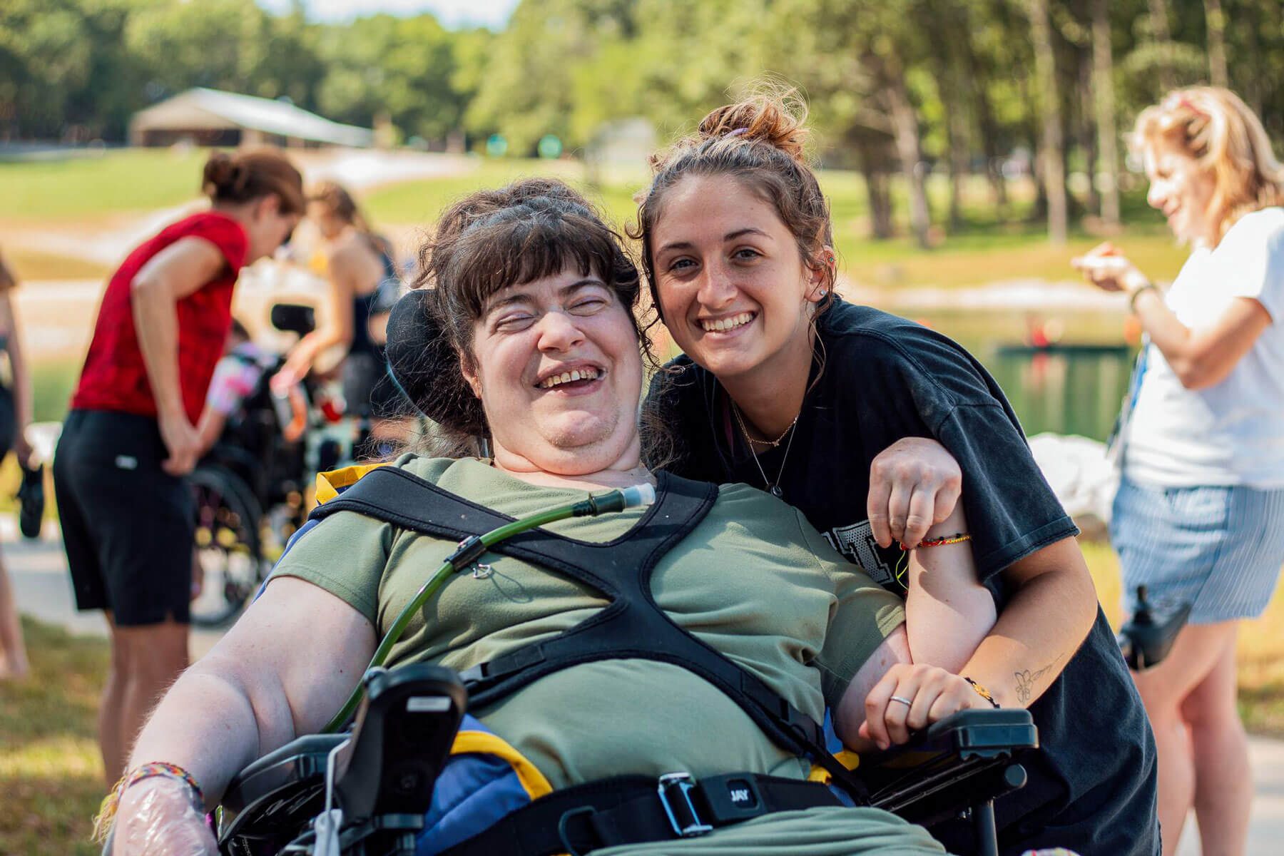 Camp counselor smiling with a camper in a wheelchair with trees and lake in the background